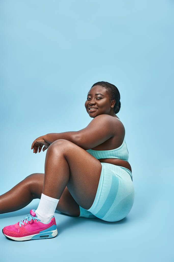 cheerful plus size african american woman sitting in blue sportswear on matching backdrop, sport - Photo, Image