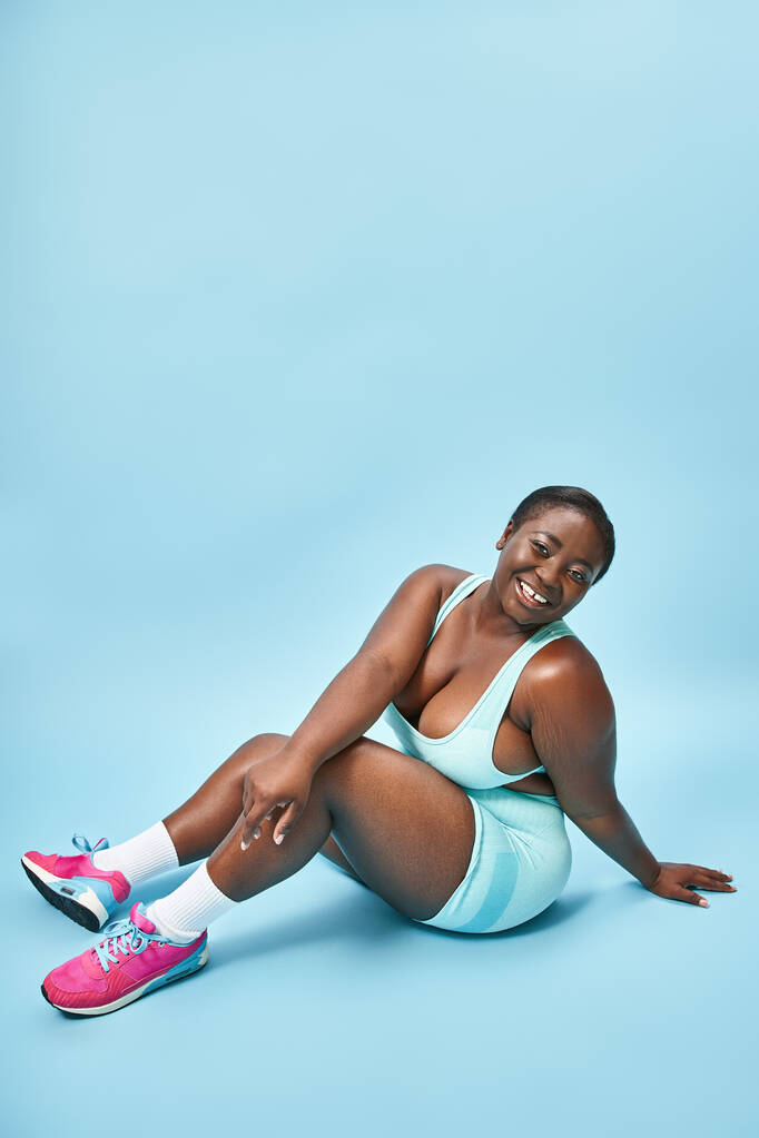 gelukkig plus size Afrikaans amerikaanse vrouw zitten in blauw sportkleding op bijpassende achtergrond, sport - Foto, afbeelding
