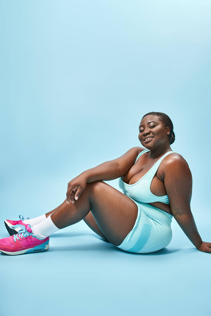 χαμογελαστός συν μέγεθος αφροαμερικανή γυναίκα κάθεται σε μπλε αθλητικά ρούχα για το ταίριασμα φόντο, τον αθλητισμό - Φωτογραφία, εικόνα