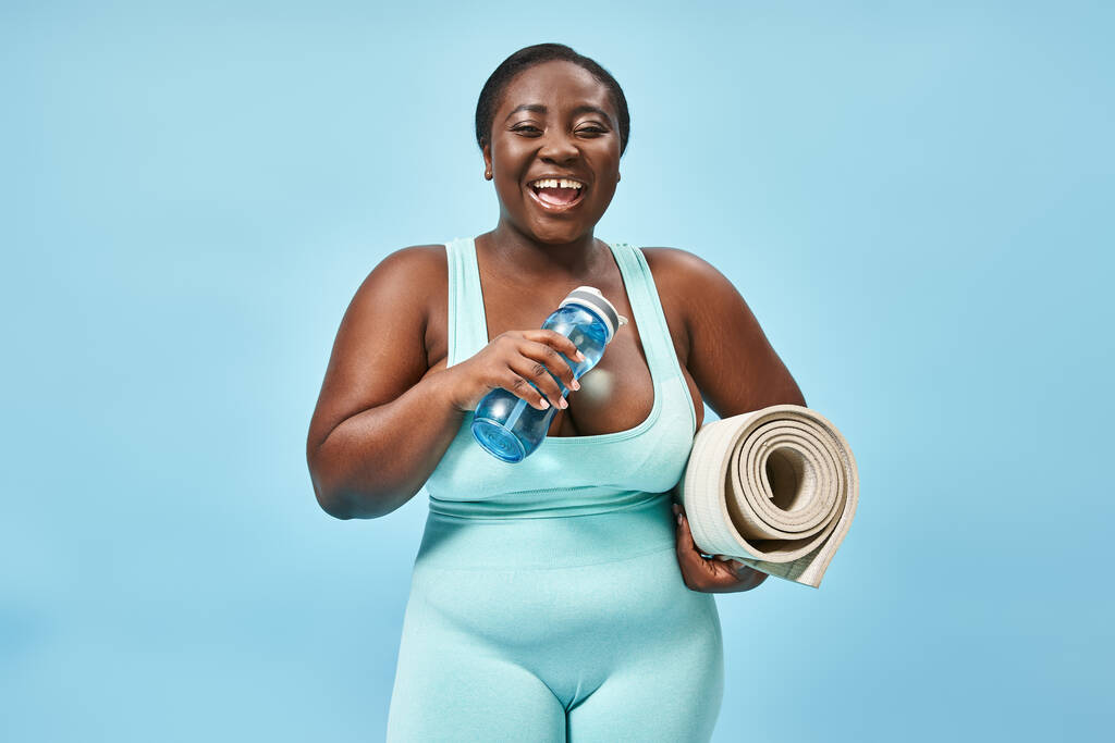 Χαμογελώντας συν μέγεθος αφροαμερικάνικη γυναίκα στέκεται με fitness mat και νερό μπουκάλι στο μπλε - Φωτογραφία, εικόνα