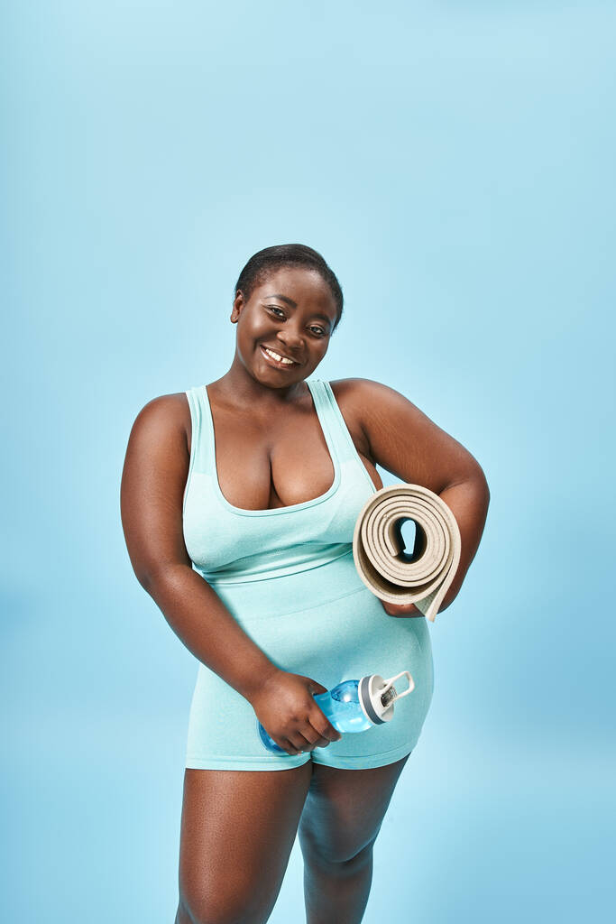 vrolijk plus size Afrikaans amerikaanse vrouw staan met fitness mat en water fles op blauw - Foto, afbeelding