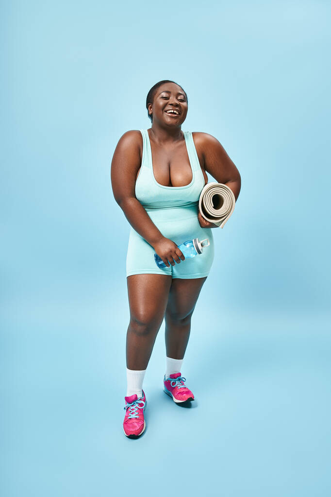 Sonriendo más tamaño mujer afroamericana de pie con tapete de fitness y botella de agua sobre fondo azul - Foto, imagen