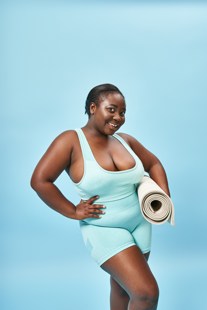 heureux plus la taille femme afro-américaine debout avec tapis de fitness sur fond bleu, sport - Photo, image