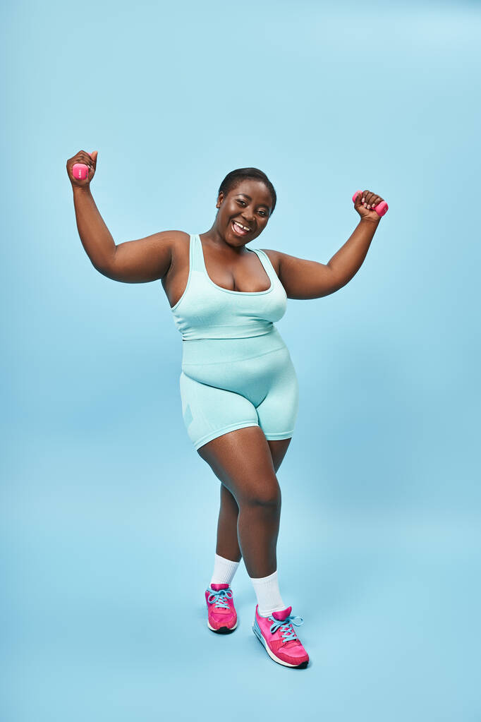 boldog plusz méret nő kék sportruházat súlyzókkal élvezi a munkáját a megfelelő háttér - Fotó, kép