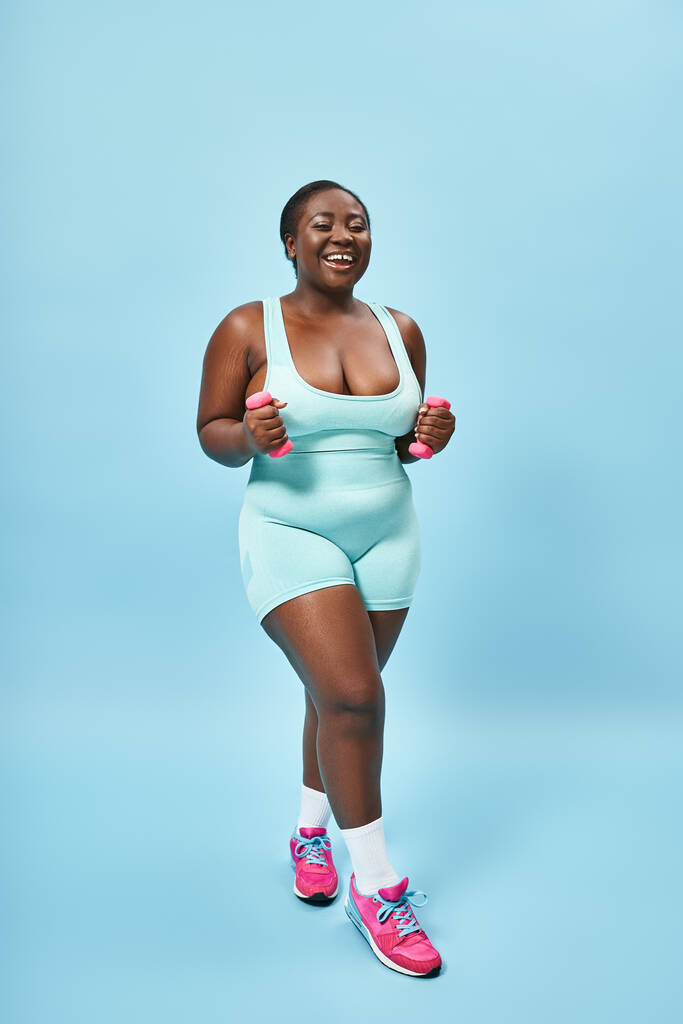 cheerful plus size woman in blue active wear exercising with dumbbells on matching background - Photo, Image