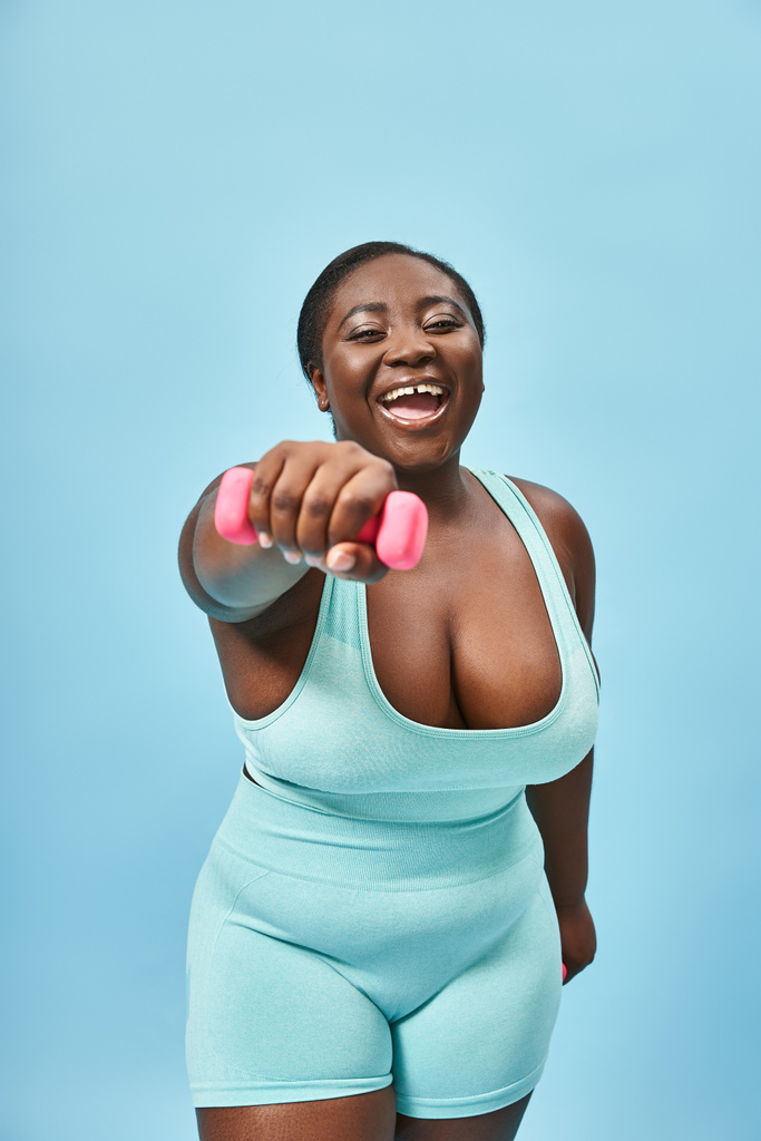joyful plus size woman in blue active wear exercising with dumbbell on matching background - Photo, Image