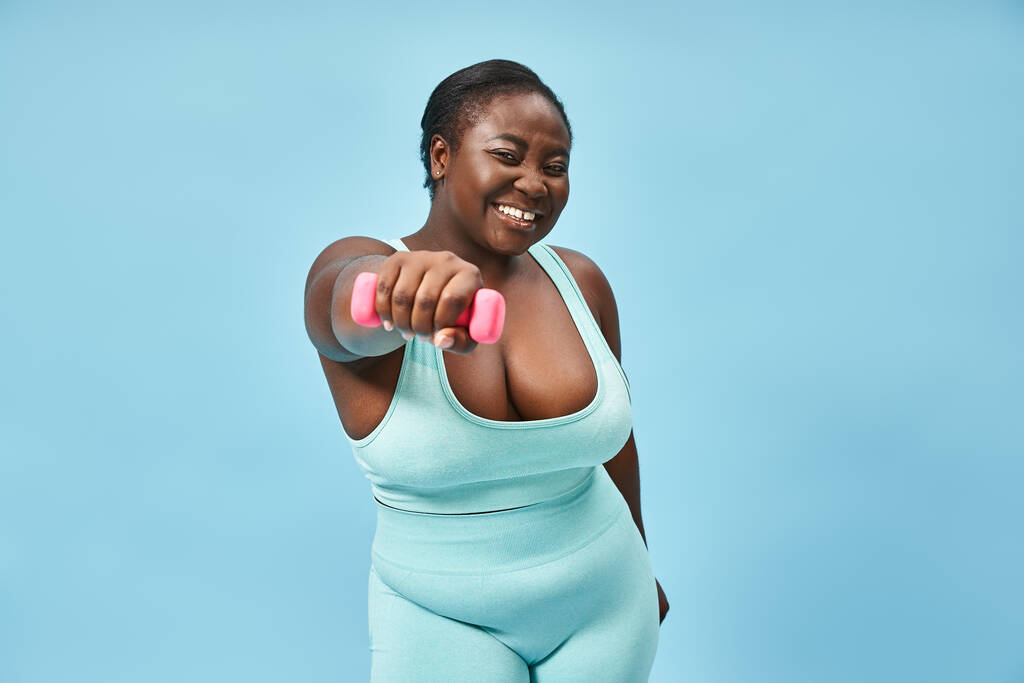 joyful plus size sportswoman in blue active wear exercising with dumbbell on matching background - Photo, Image