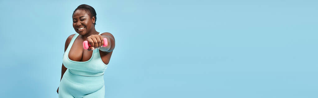gioioso plus size donna in blu attivo usura esercizio con manubri su sfondo corrispondente, banner - Foto, immagini