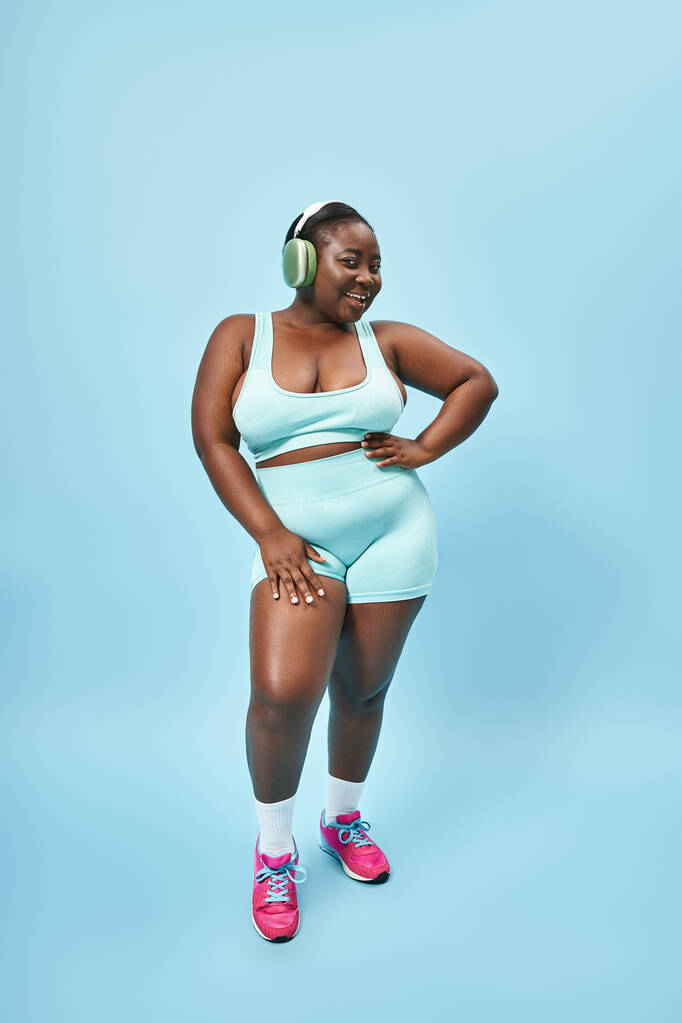 radosna plus size Afrykańska amerykanka w aktywnym ubraniu słuchanie muzyki w słuchawkach, niebieskie tło - Zdjęcie, obraz