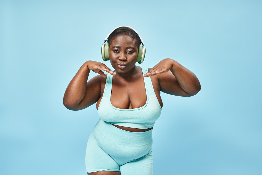 young plus size african american woman in active wear listening music in headphones, blue backdrop - Photo, Image