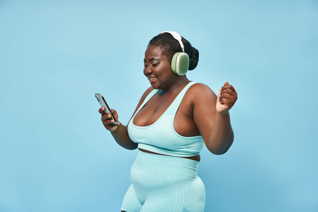 plus size Afrykańska amerykańska kobieta słuchająca muzyki w słuchawkach i trzymająca smartfona, niebieskie tło - Zdjęcie, obraz