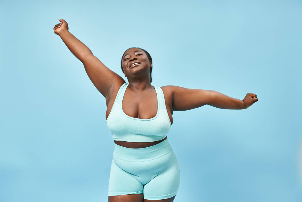 happy plus size african american woman in active wear stretching joyfully on blue background - Photo, Image