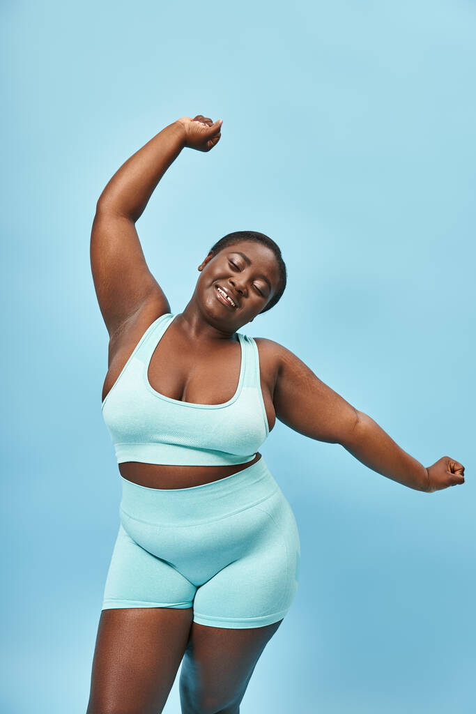 cheerful plus size african american woman in active wear stretching joyfully on blue background - Photo, Image