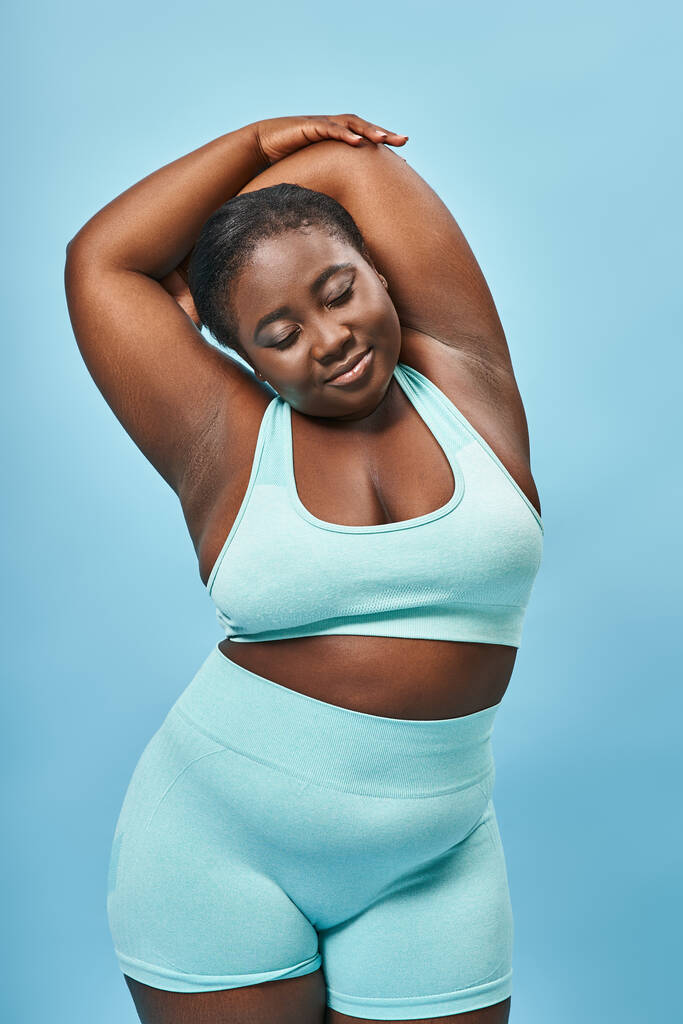zadowolony plus rozmiar Afroamerykanka kobieta w aktywnym nosić rozciągające się na niebieskim tle, ciało pozytywne - Zdjęcie, obraz