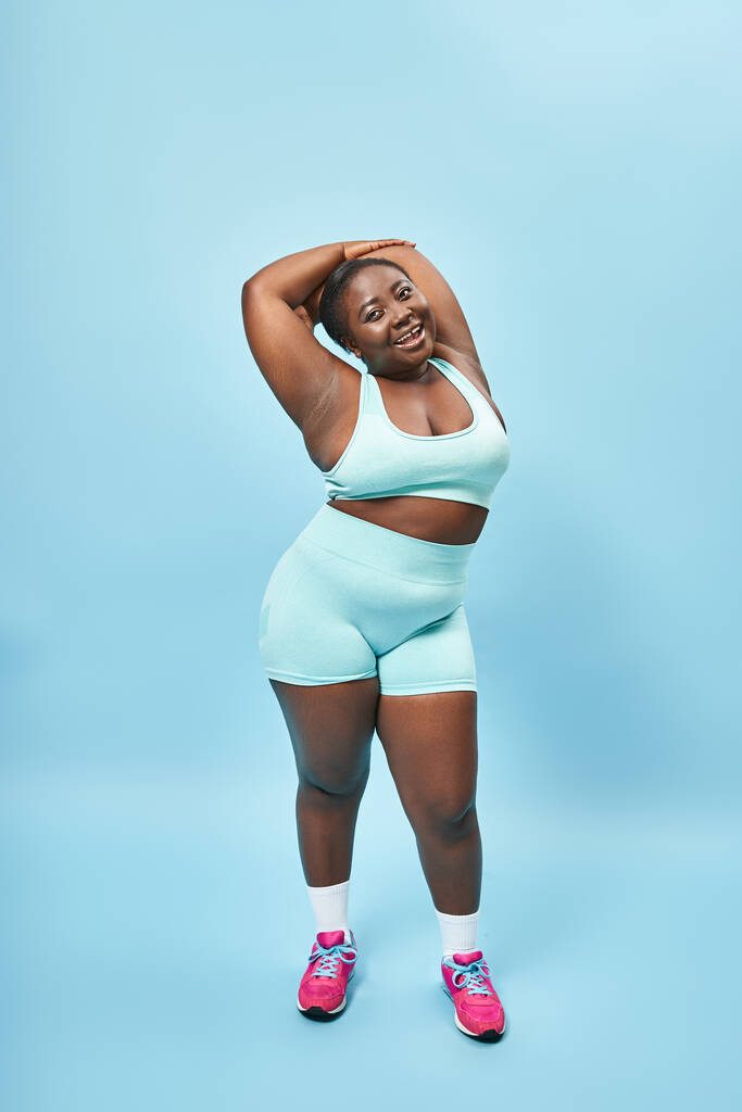 erfreut plus size afrikanisch-amerikanische Frau in aktiver Kleidung Stretching auf blauem Hintergrund, Körper positiv - Foto, Bild