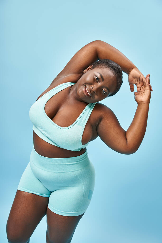 gai plus la taille femme afro-américaine en tenue active étirant sur fond bleu, corps positif - Photo, image