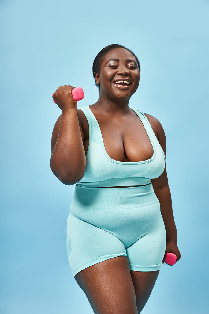θετική στο σώμα, συν μέγεθος γυναίκα σε μπλε ενεργή φθορά άσκηση με αλτήρες στο φόντο ταιριάζουν - Φωτογραφία, εικόνα
