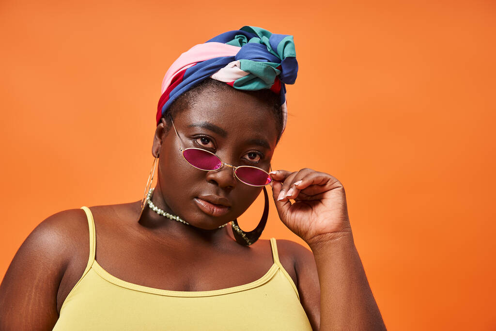 Fashion-forward en plus size Afrikaans Amerikaans model in hoofddoek en trendy zonnebril op oranje - Foto, afbeelding