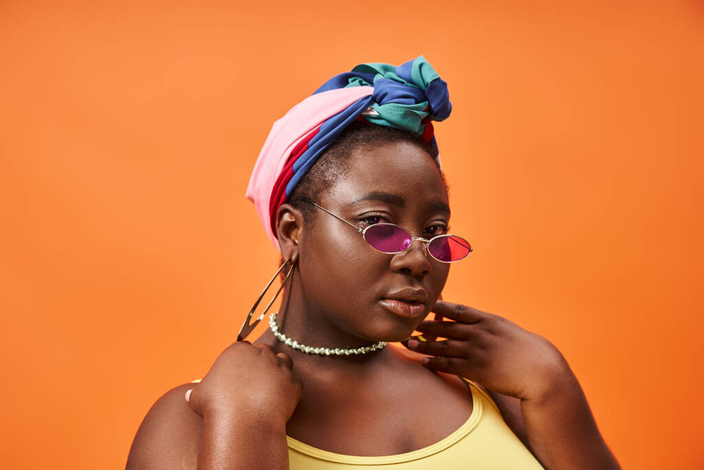 De moda más tamaño y mujer afroamericana joven en pañuelo para la cabeza y gafas de sol de moda en naranja - Foto, Imagen
