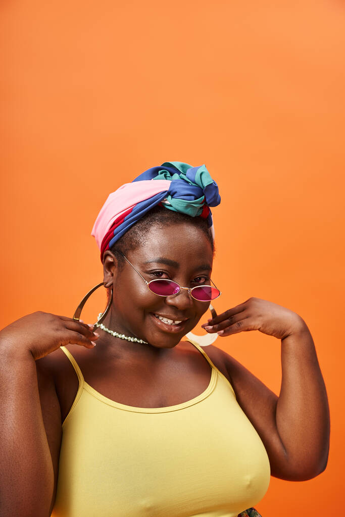 オレンジのヘッドカーフとトレンディなサングラスでファッショナブルなプラスサイズと幸せなアフリカ系アメリカ人女性 - 写真・画像