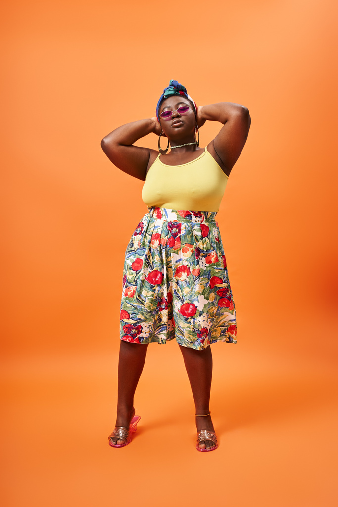 mujer afroamericana con estilo más tamaño en traje floral y gafas de sol de moda sobre fondo naranja - Foto, Imagen