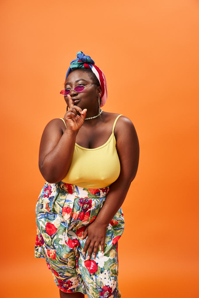плюс размер африканский американец женщина в цветочном костюме и солнцезащитные очки показывая знак молчания на оранжевом - Фото, изображение
