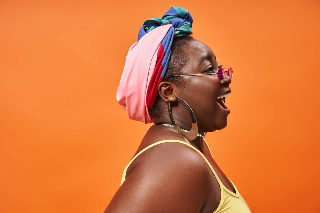 vista lateral de mujer afroamericana de talla grande en pañuelo para la cabeza y gafas de sol de moda sobre fondo naranja - Foto, imagen
