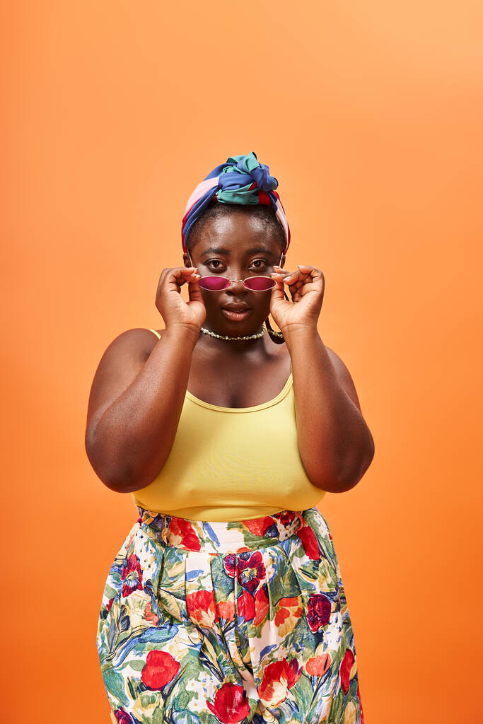 плюс размер африканская американка в цветочном костюме носить стильные солнцезащитные очки на оранжевом фоне - Фото, изображение