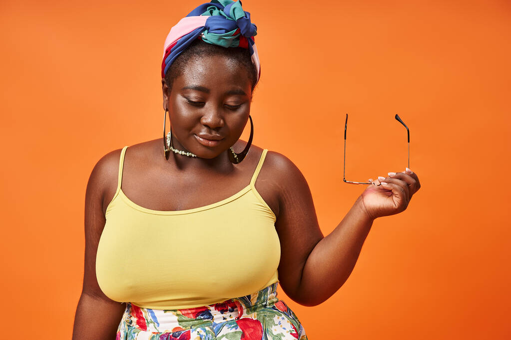 plus size african american woman in floral outfit holding trendy sunglasses on orange background - Photo, Image