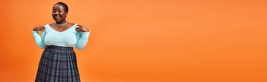 felice plus size donna africana americana in gonna a quadri e maniche lunghe blu in posa su arancione, banner - Foto, immagini