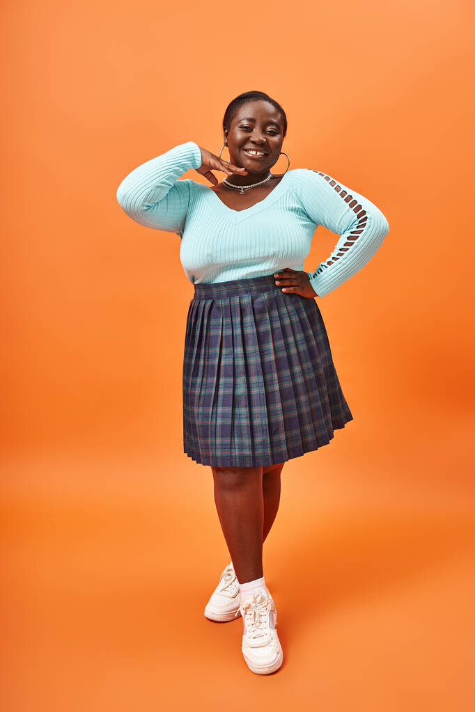 boldog plusz méret afro-amerikai nő kockás szoknya és hosszú ujjú mosolygós narancs háttér - Fotó, kép