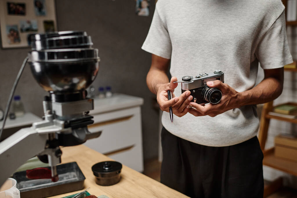 recortado de hombre negro Sujetando intencionadamente una cámara analógica y un bolígrafo mientras está de pie en un laboratorio fotográfico - Foto, imagen