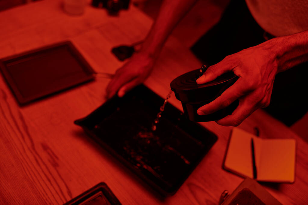 Overhead-Ansicht des Fotografen gießt Entwickler in Tablett zur Filmbearbeitung in roter Dunkelkammer - Foto, Bild