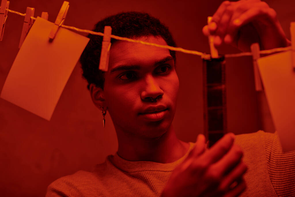 joven afroamericano hombre cuelga tira de película recién desarrollado en un cuarto oscuro iluminado por el rojo, atemporal - Foto, Imagen