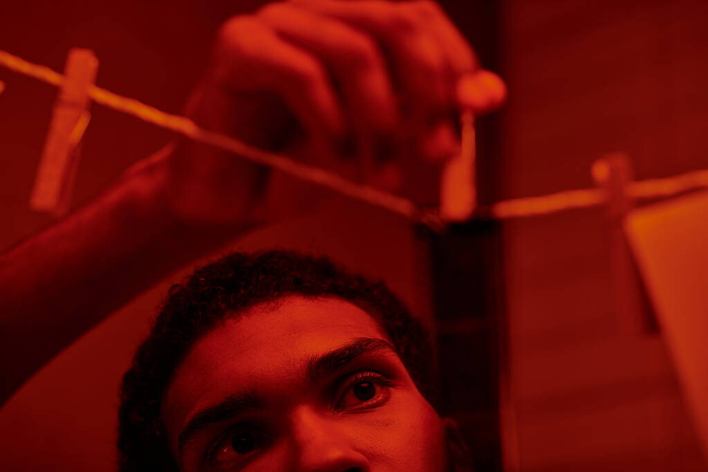joven afroamericano hombre cuelga recién desarrollado tira de película en un cuarto oscuro de luz roja, nostalgia - Foto, imagen