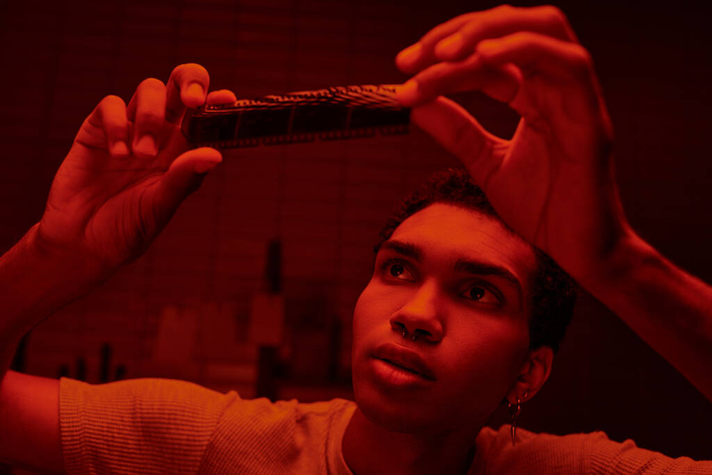 fokussierter afrikanisch-amerikanischer Fotograf beim Betrachten eines entwickelten Filmstreifens in einer rotbeleuchteten Dunkelkammer - Foto, Bild