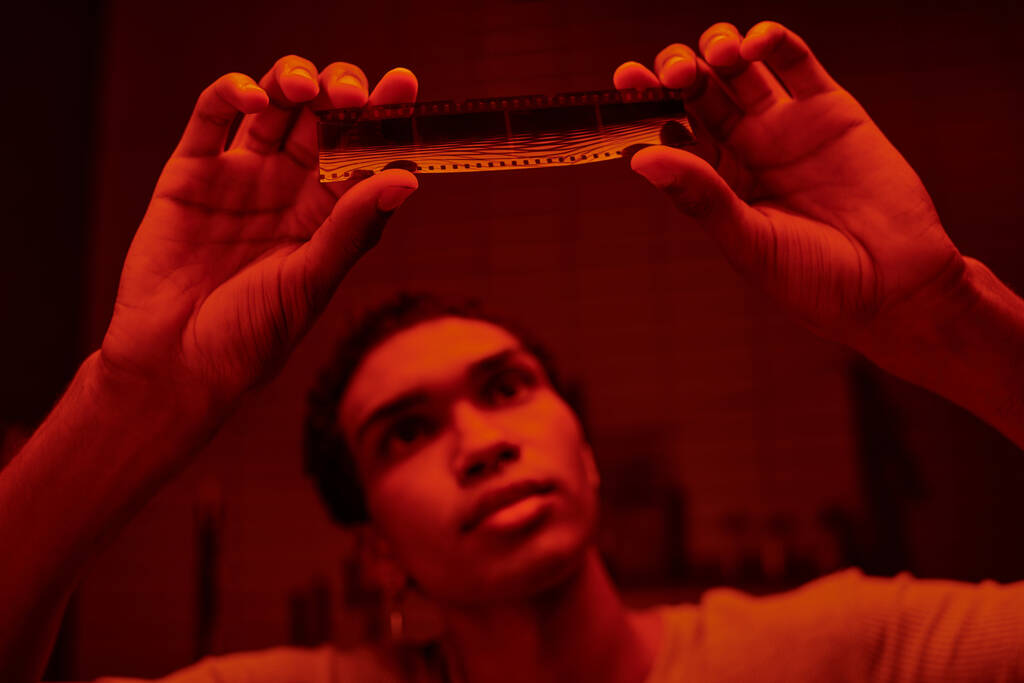 fotógrafo afroamericano enfocado examinar desarrollado tira de película en un cuarto oscuro con luz roja - Foto, imagen