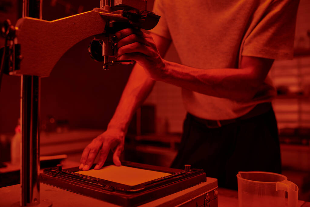 cropped photographer focuses on delicate process of enlarging film in darkroom with red light - Photo, Image