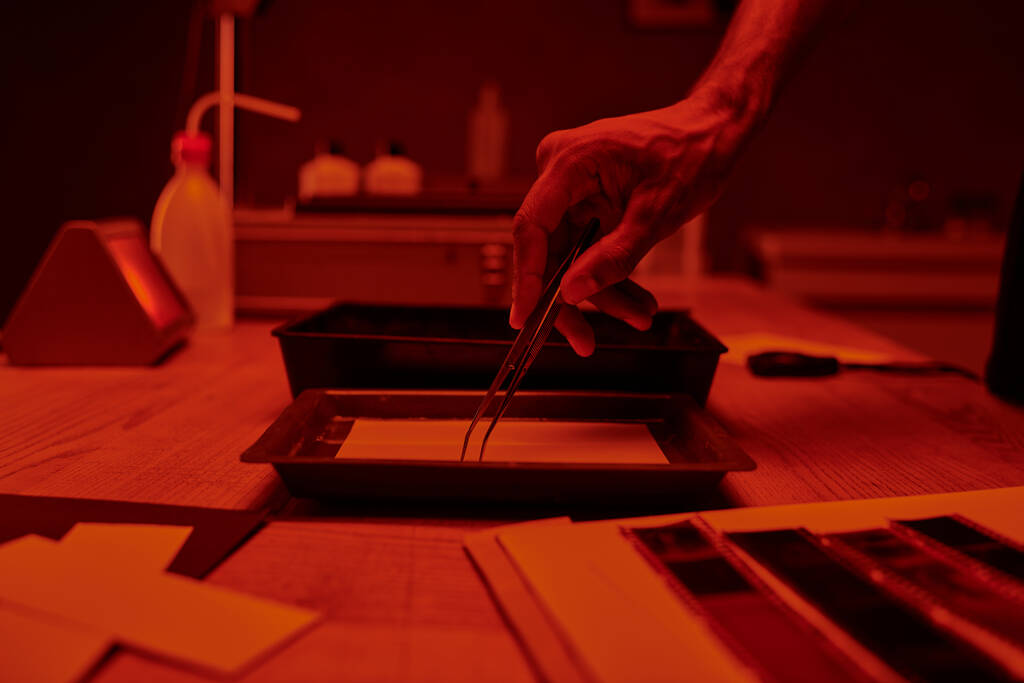 cropped view of photographer delicately handling film with tweezers under red light in darkroom - Photo, Image