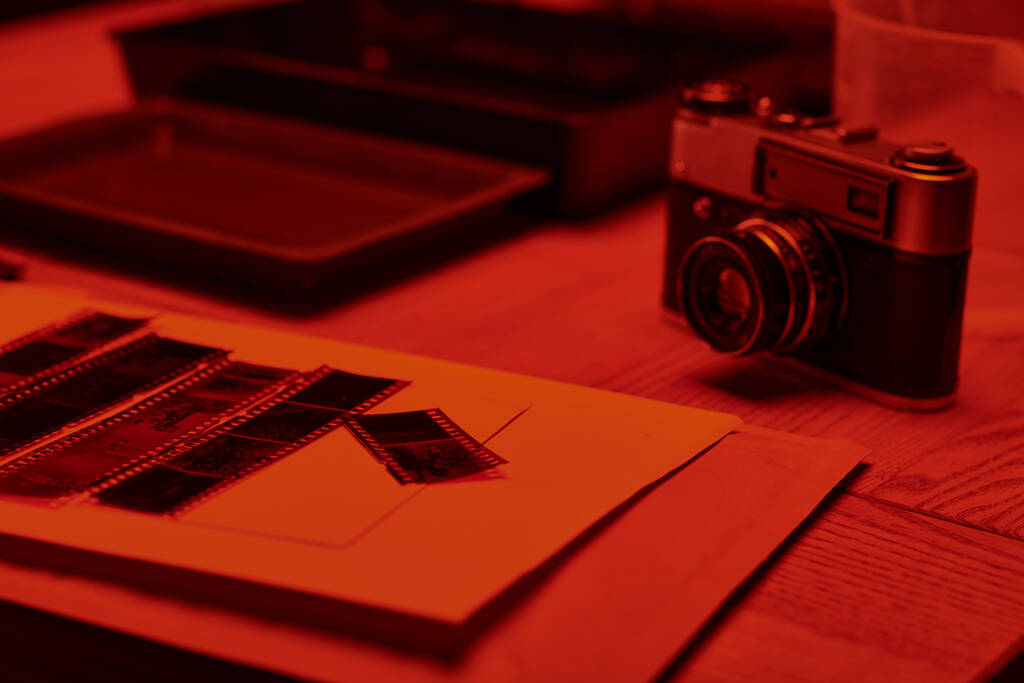 Стіл з аналоговою камерою і смужками плівки під сяйвом червоного світла в темній кімнаті, поза часом - Фото, зображення