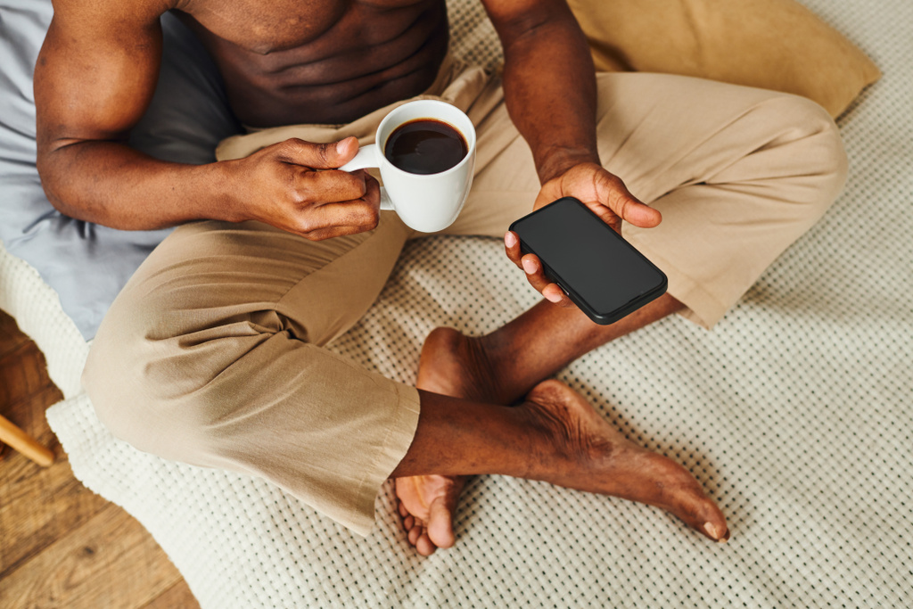 Afrikkalainen amerikkalainen mies pyjamassa housut istuu sängyllä kahvia ja älypuhelin tyhjällä näytöllä - Valokuva, kuva