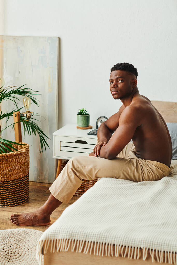 σκεπτόμενος αφρικάνος Αμερικανός χωρίς πουκάμισο άντρας με παντελόνι πιτζάμας κάθεται και κοιτάζει την κάμερα στην κρεβατοκάμαρα - Φωτογραφία, εικόνα