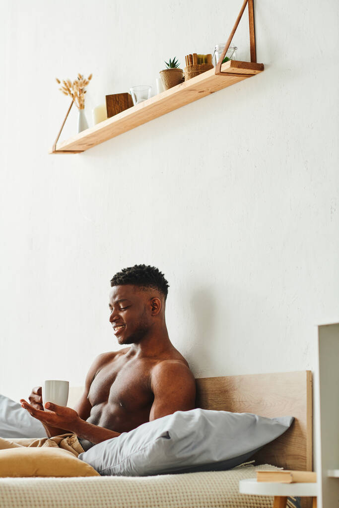 радісний афроамериканський чоловік з сорочкою м'язистий торс, що сидить з чашкою кави в спальні - Фото, зображення