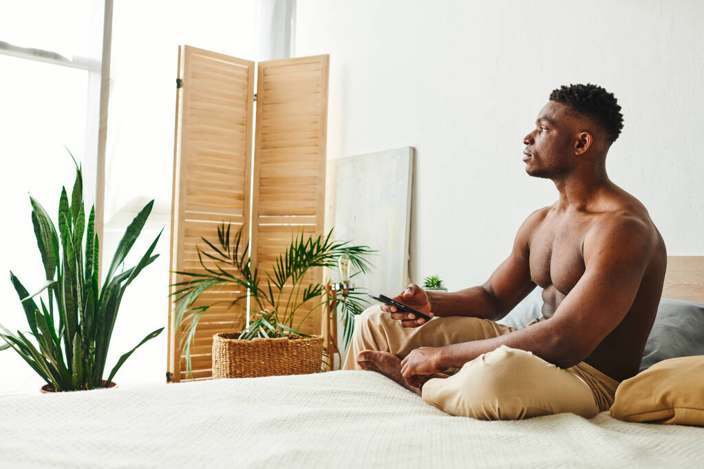 夢見る アフリカ系アメリカ人男性 パジャマパンツ スマートフォンでベッドに座って離れて見る - 写真・画像