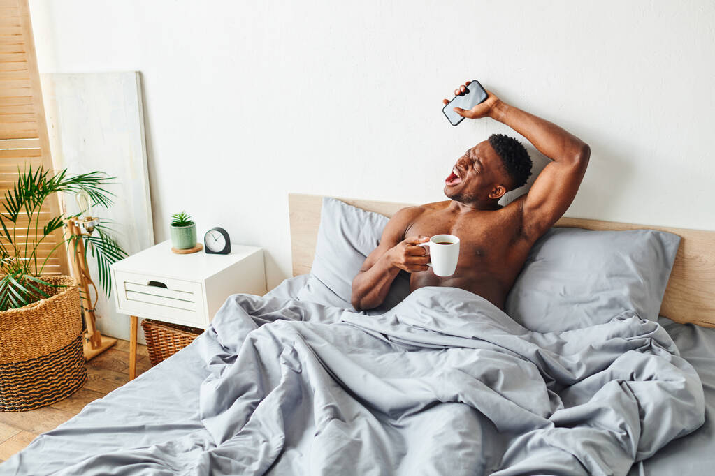 young african american guy with coffee cup and smartphone yawning and stretching waking up on bed - Photo, Image
