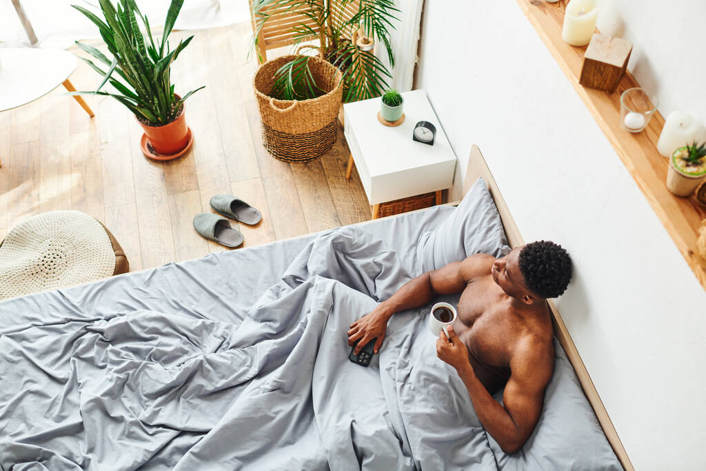 вид сверху на мускулистого африканского парня с чашкой кофе и смартфоном, смотрящего на кровать - Фото, изображение