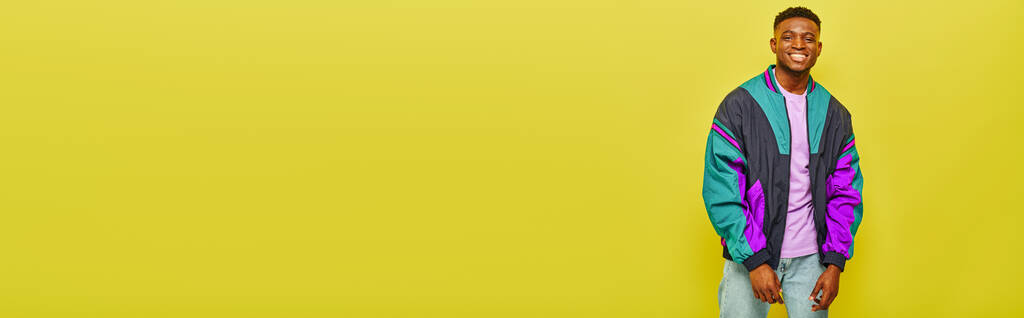 Renkli rüzgarlık giymiş neşeli Afrikalı Amerikalı adam sarı arka planda gülümsüyor ve poz veriyor. - Fotoğraf, Görsel