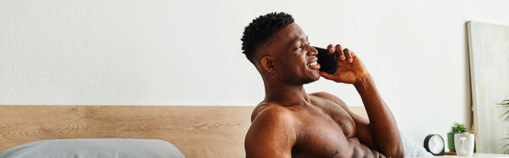 Gülümseyen kaslı Afrikalı Amerikalı adam cep telefonuyla konuşuyor modern rahat yatak odasında, pankartta - Fotoğraf, Görsel