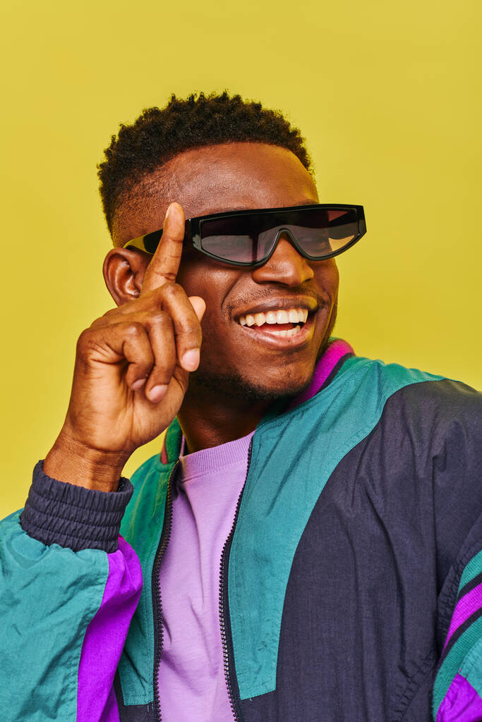 Χαρούμενος Αφροαμερικάνος με αντιανεμικό μπουφάν που προσαρμόζει γυαλιά ηλίου και κοιτάζει αλλού με κίτρινο. - Φωτογραφία, εικόνα