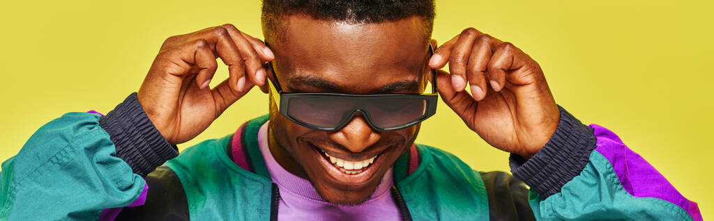 trendy Afrikaan in felle jas die zijn zonnebril aanpast en glimlacht op een gele achtergrond, spandoek - Foto, afbeelding